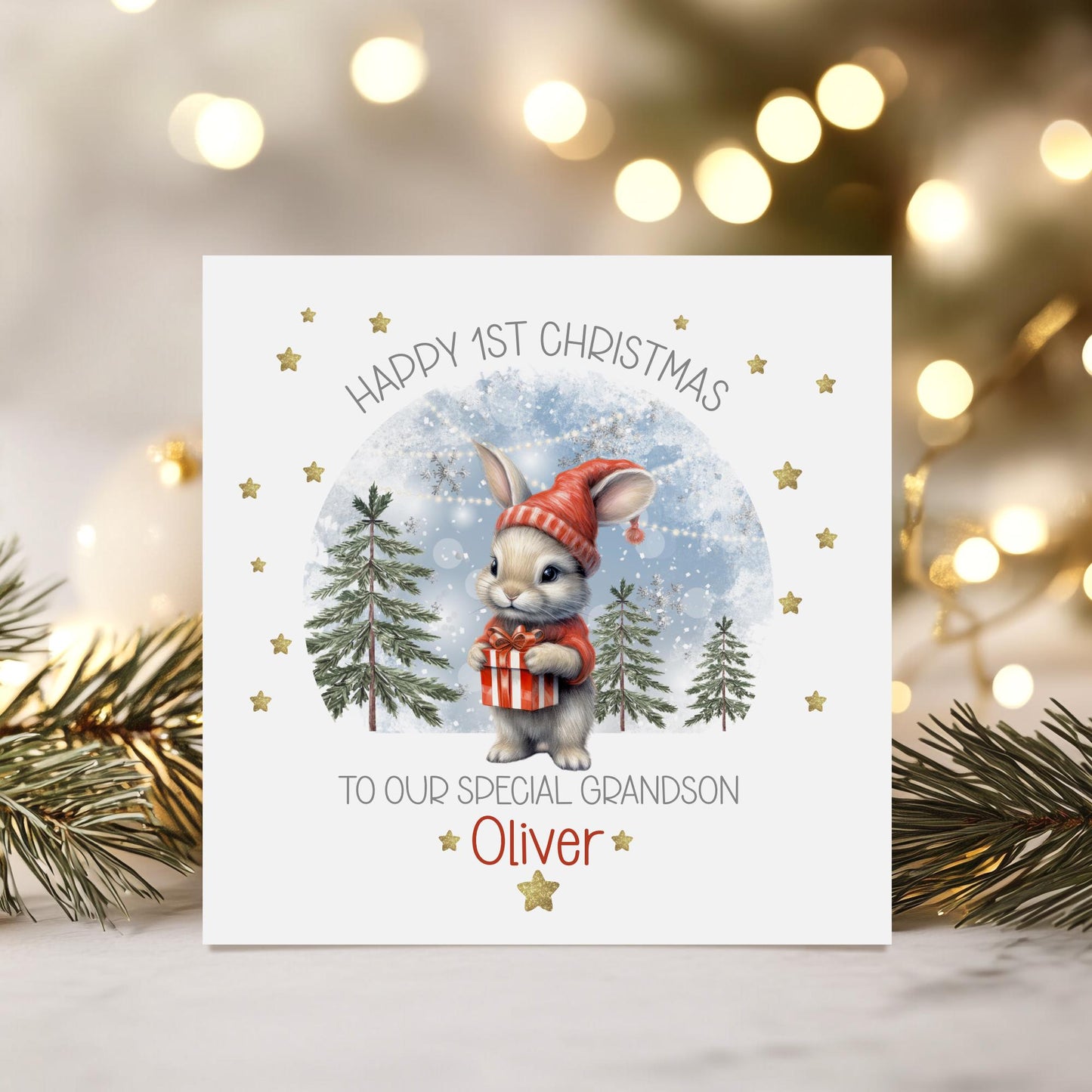 Personalised baby’s 1st Christmas card for Boys & girls featuring a cute bunny in a red hat holding a present, snowy woodland background with fir trees and gold stars. Printed on premium 300gsm card.