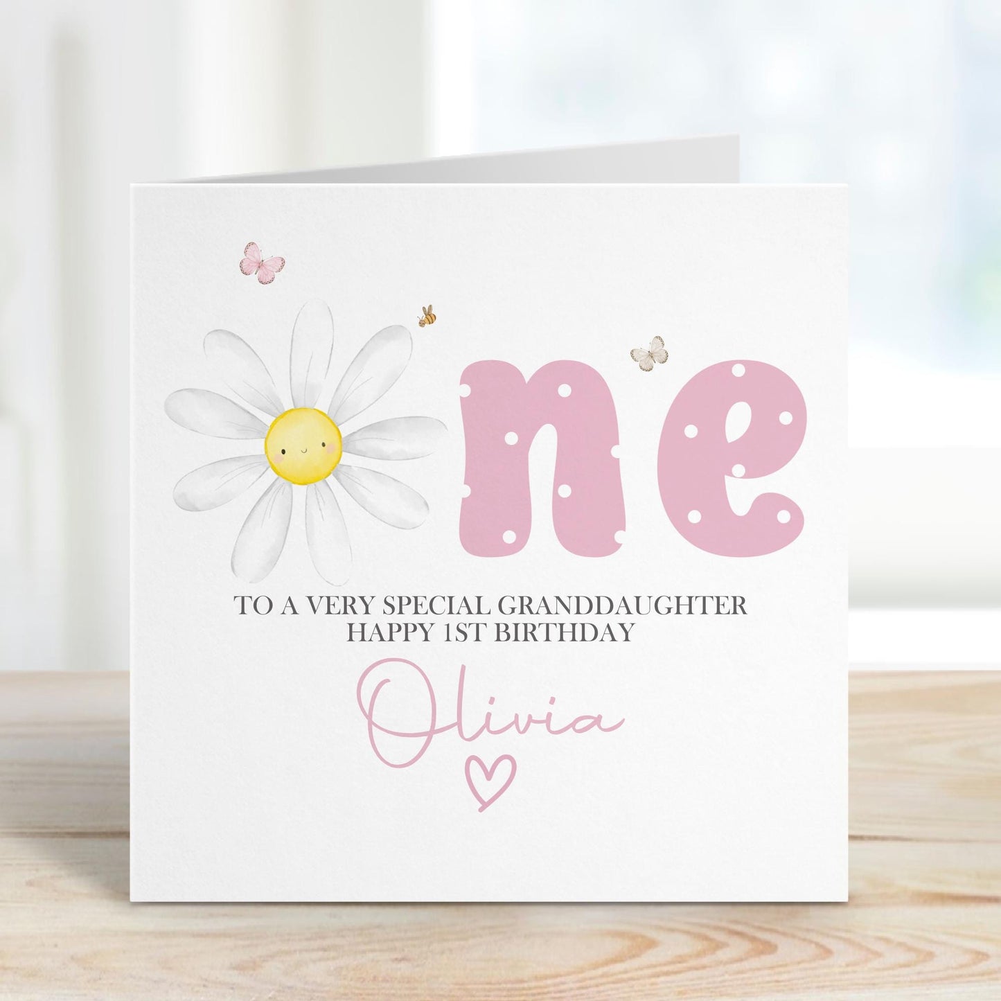 Luxury white linen, lightly textured 1st birthday card featuring a watercolor daisy & the one word ONE in pink polka dot & pretty butterflys & a bee & the wording To a very soecial granddaughter, happy 1st birthday, personalised with the childs name a little heart. Card measures 6 x 6 inches.