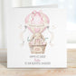 Square personalised Easter greeting card featuring an illustrated bunny sitting in a wicker basket hot air balloon in soft pink pastel tones. The design includes Easter eggs, ribbon details and a banner reading ‘Happy Easter’. The card has space for personalised text for name, year and relation.