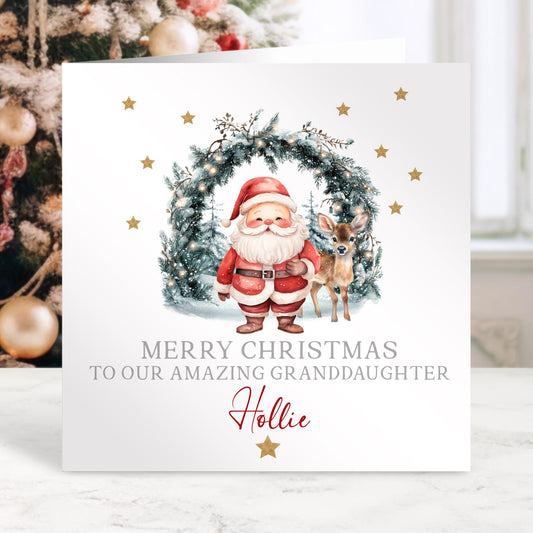 Personalised Christmas card featuring Santa and a reindeer standing in front of a festive wreath with twinkling lights, gold stars, and the message ‘Merry Christmas to our amazing granddaughter Hollie’ printed on the front. High-quality 300gsm card.
