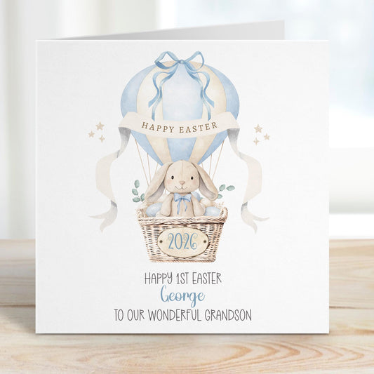Square personalised Easter greeting card featuring a cute illustrated bunny sitting in a wicker hot air balloon basket surrounded by pastel Easter eggs. The balloon is decorated in soft blue tones with a ribbon and a banner reading ‘Happy Easter’. The design includes space for personalised text for name, year and relation.