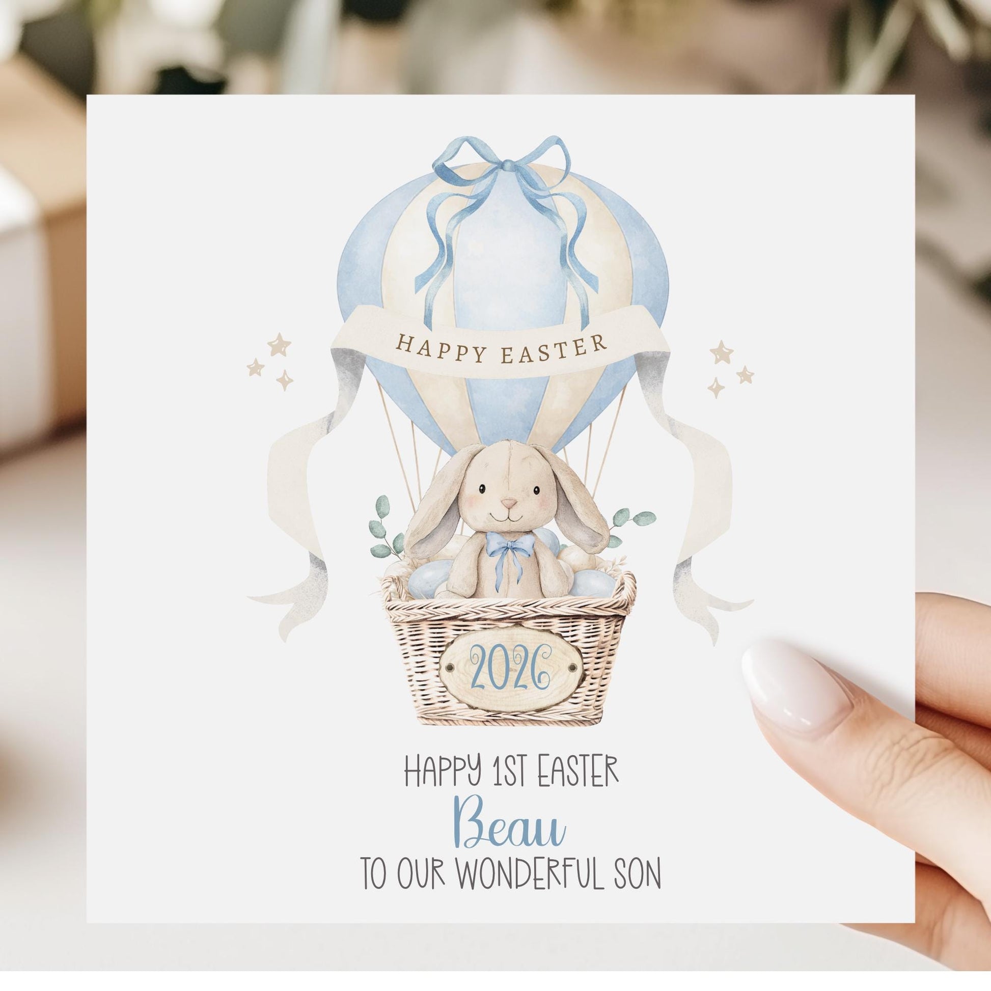 Square personalised Easter greeting card featuring a cute illustrated bunny sitting in a wicker hot air balloon basket surrounded by pastel Easter eggs. The balloon is decorated in soft blue tones with a ribbon and a banner reading ‘Happy Easter’. The design includes space for personalised text for name, year and relation.