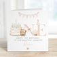 Square personalised first birthday greeting card featuring soft pastel illustrations including a teddy bear, birthday cake with candle, bunting, flowers and a gift basket. The design displays the word ‘ONE’ with decorative details and space for personalised name and relation text.