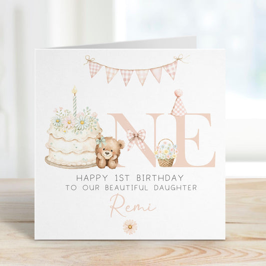Square personalised first birthday greeting card featuring soft pastel illustrations including a teddy bear, birthday cake with candle, bunting, flowers and a gift basket. The design displays the word ‘ONE’ with decorative details and space for personalised name and relation text.