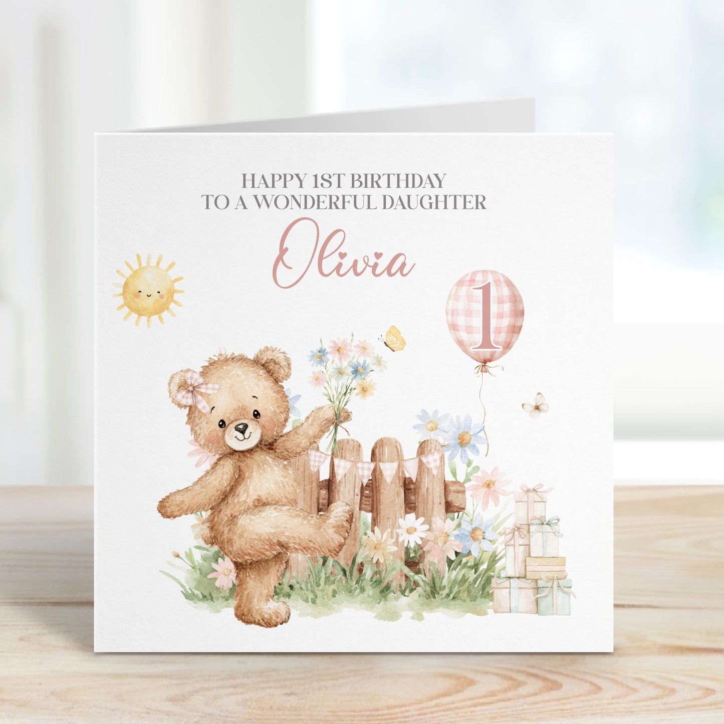 Square personalised first birthday greeting card featuring a cute illustrated teddy bear standing in a garden scene with flowers, a wooden fence, sunshine, gifts and a balloon showing the number one. The card includes personalised text for a child’s name and relation, with a soft pastel colour theme.
