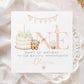 Square personalised first birthday greeting card featuring soft pastel illustrations including a teddy bear, birthday cake with candle, bunting, flowers and a gift basket. The design displays the word ‘ONE’ with decorative details and space for personalised name and relation text.