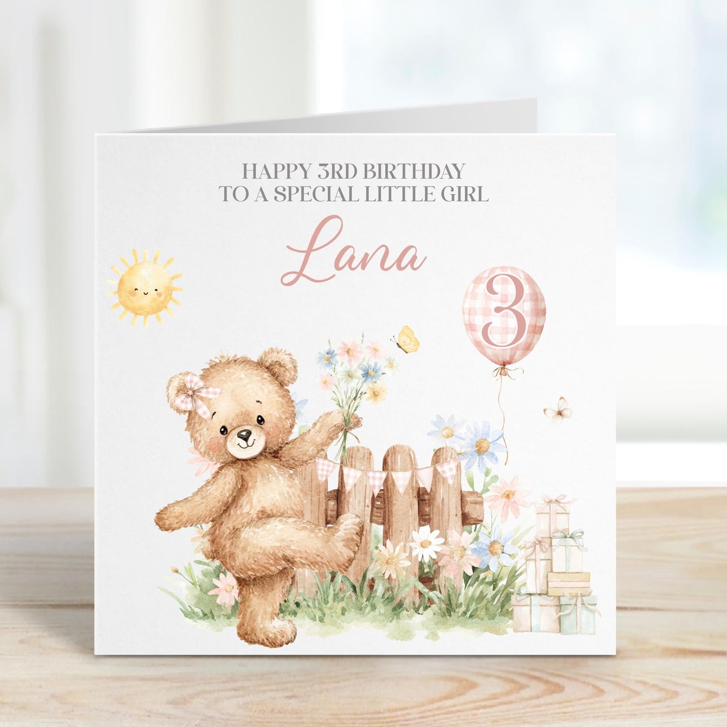 Square personalised third birthday greeting card featuring a cute illustrated teddy bear standing in a garden scene with flowers, a wooden fence, sunshine, gifts and a balloon showing the number three. The card includes personalised text for a child’s name and relation, with a soft pastel colour theme.