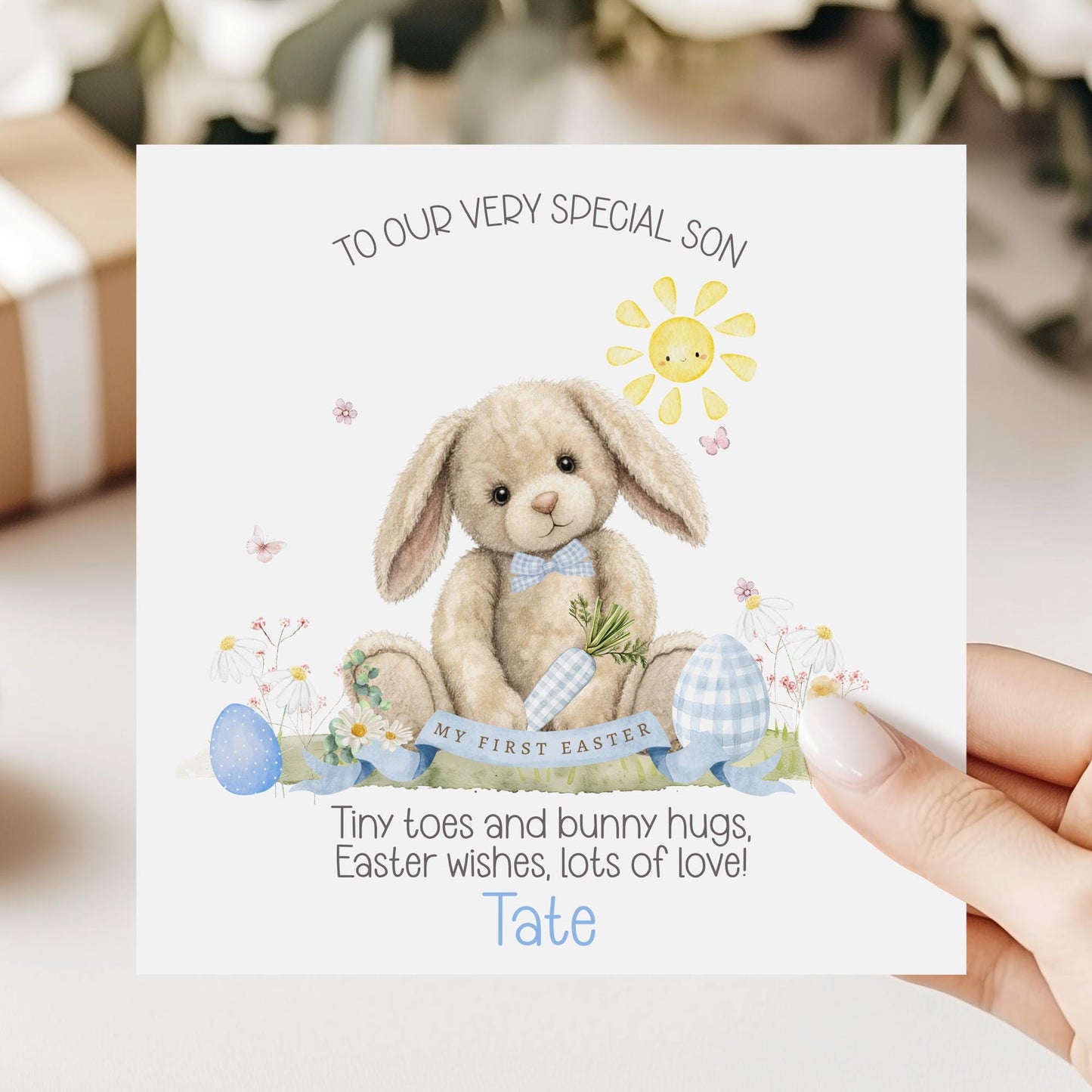 Personalised first Easter card for a grandson featuring a soft illustrated bunny holding a carrot, surrounded by pastel Easter eggs, flowers and a smiling sun, with “My First Easter” banner and space for a personalised name.