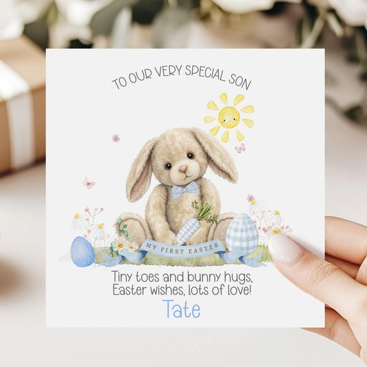 Personalised first Easter card for a grandson featuring a soft illustrated bunny holding a carrot, surrounded by pastel Easter eggs, flowers and a smiling sun, with “My First Easter” banner and space for a personalised name.