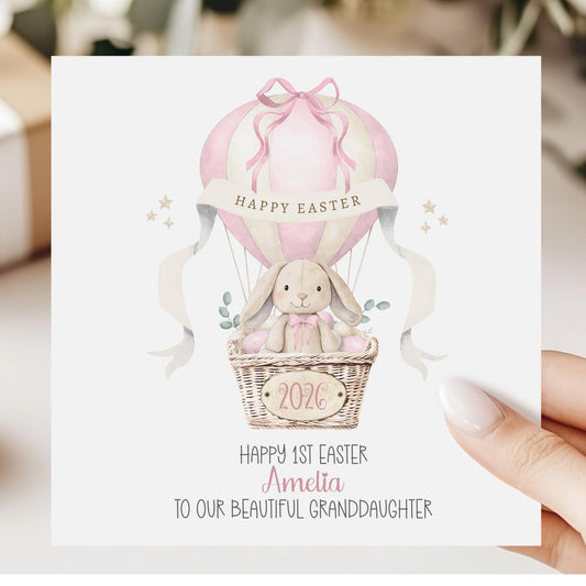 Square personalised Easter greeting card featuring an illustrated bunny sitting in a wicker basket hot air balloon in soft pink pastel tones. The design includes Easter eggs, ribbon details and a banner reading ‘Happy Easter’. The card has space for personalised text for name, year and relation.