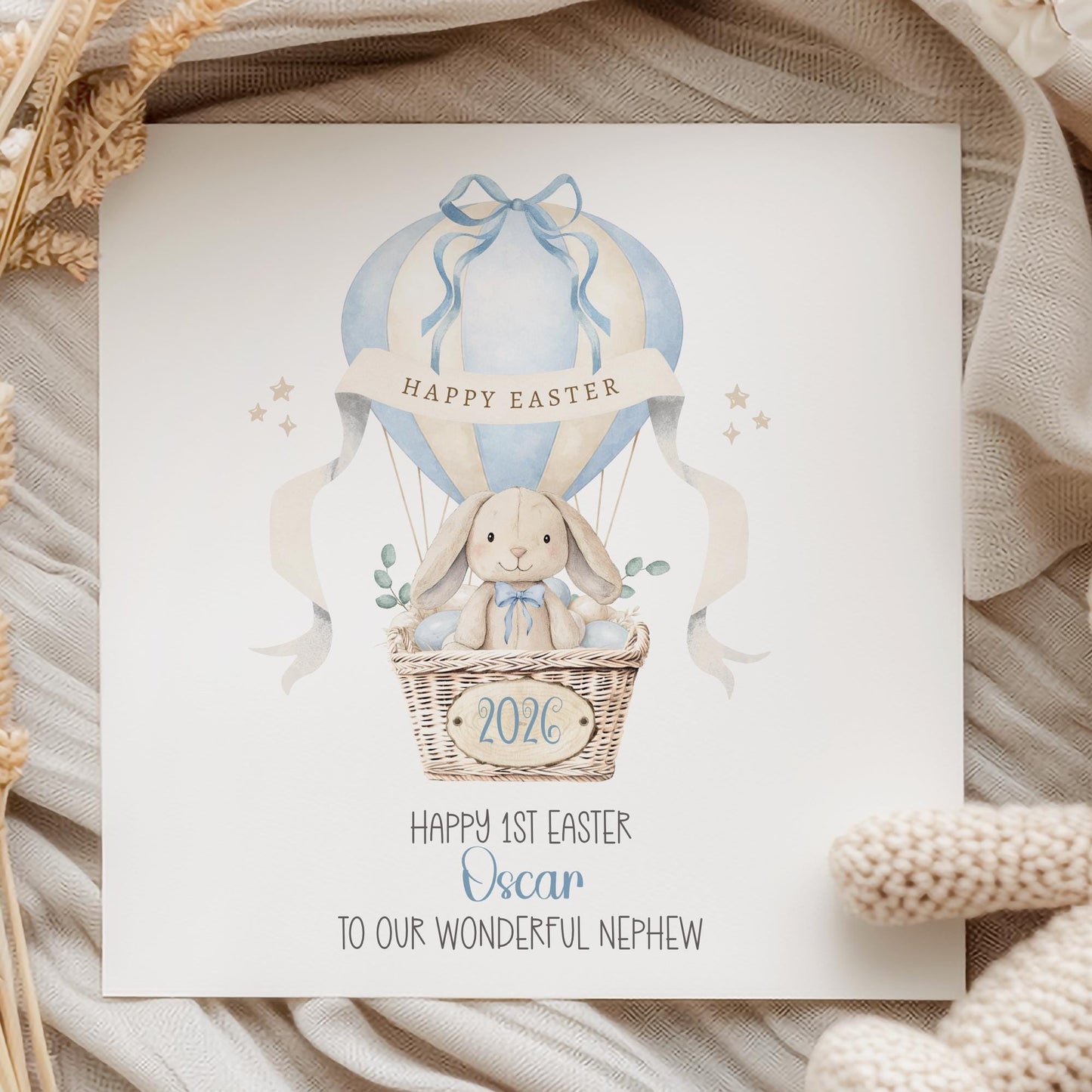Square personalised Easter greeting card featuring a cute illustrated bunny sitting in a wicker hot air balloon basket surrounded by pastel Easter eggs. The balloon is decorated in soft blue tones with a ribbon and a banner reading ‘Happy Easter’. The design includes space for personalised text for name, year and relation.