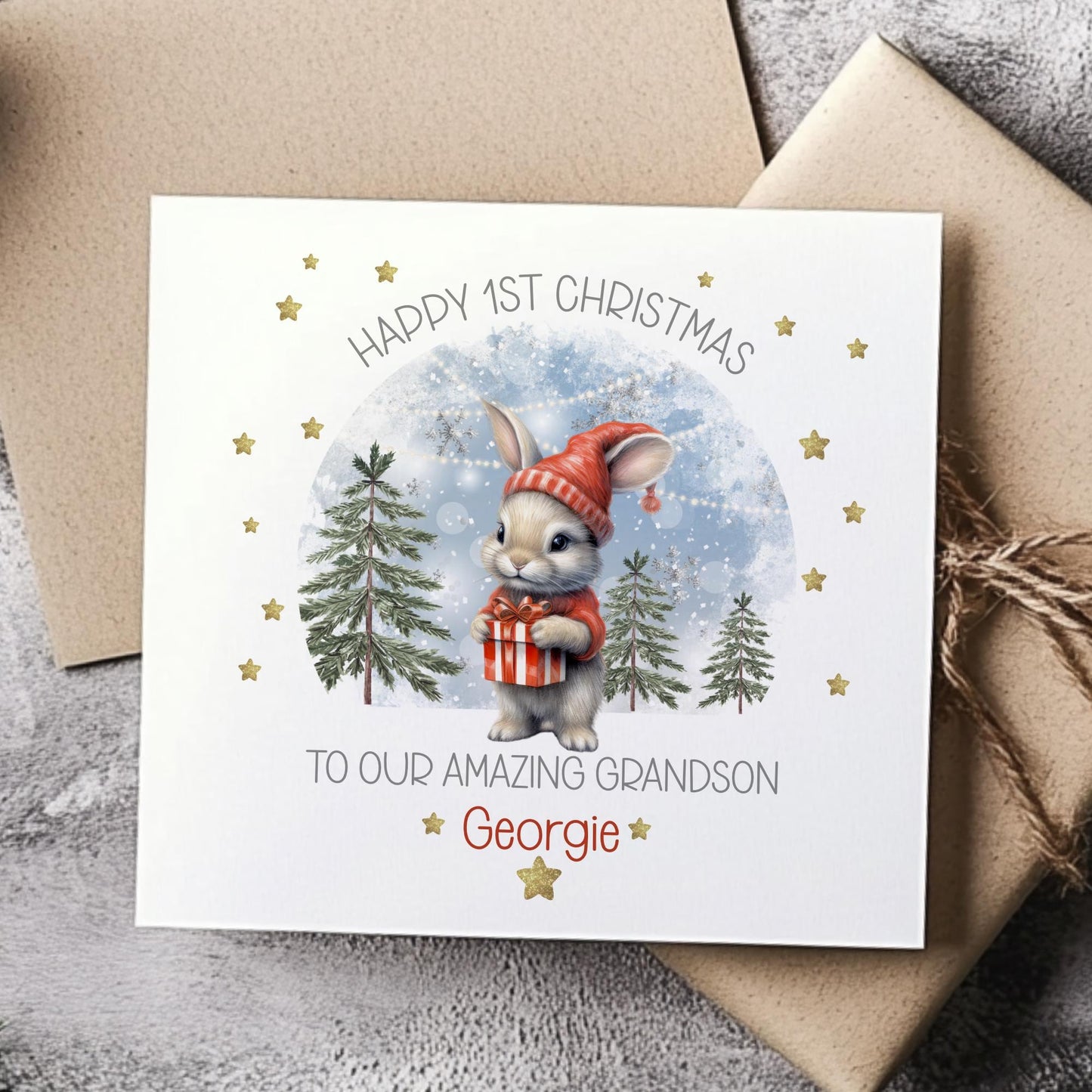 Personalised baby’s 1st Christmas card for Boys & girls featuring a cute bunny in a red hat holding a present, snowy woodland background with fir trees and gold stars. Printed on premium 300gsm card.
