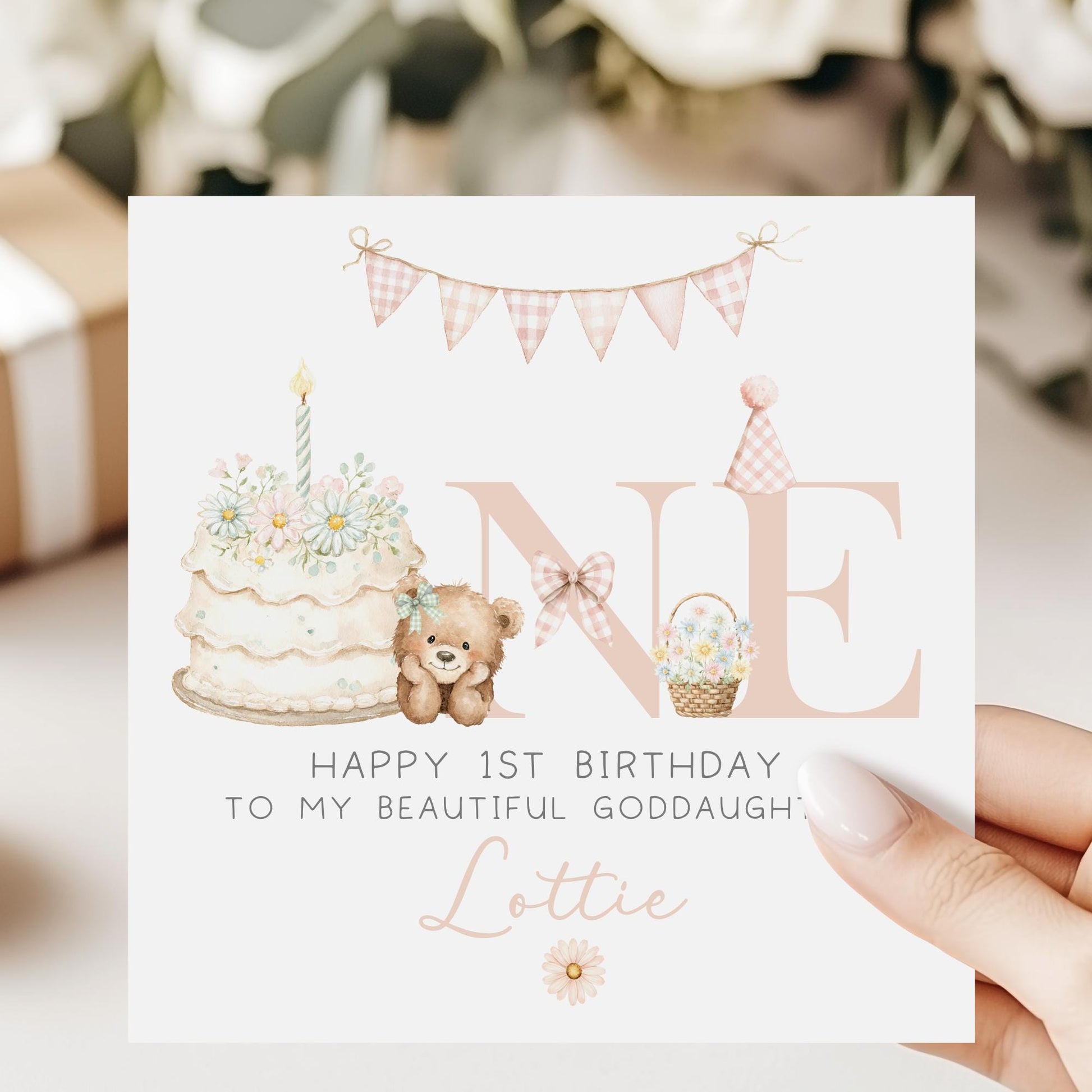 Square personalised first birthday greeting card featuring soft pastel illustrations including a teddy bear, birthday cake with candle, bunting, flowers and a gift basket. The design displays the word ‘ONE’ with decorative details and space for personalised name and relation text.