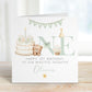 Square personalised first birthday greeting card featuring soft pastel illustrations including a teddy bear, birthday cake with candle, bunting, flowers and a gift basket. The design displays the word ‘ONE’ with decorative details and space for personalised name and relation text.