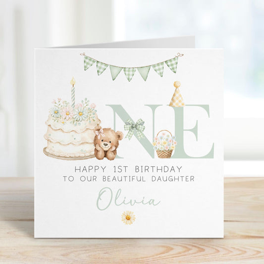 Square personalised first birthday greeting card featuring soft pastel illustrations including a teddy bear, birthday cake with candle, bunting, flowers and a gift basket. The design displays the word ‘ONE’ with decorative details and space for personalised name and relation text.