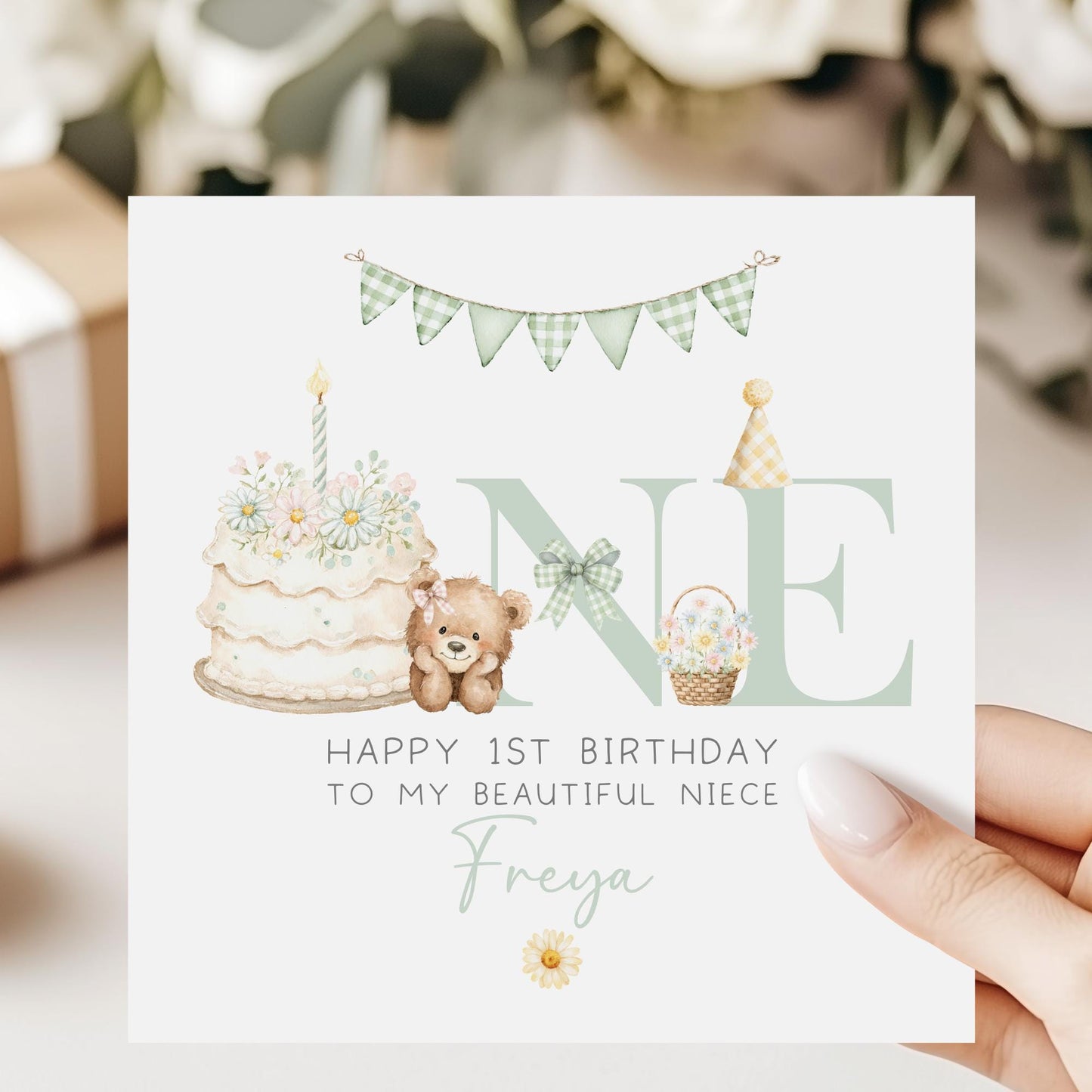 Square personalised first birthday greeting card featuring soft pastel illustrations including a teddy bear, birthday cake with candle, bunting, flowers and a gift basket. The design displays the word ‘ONE’ with decorative details and space for personalised name and relation text.