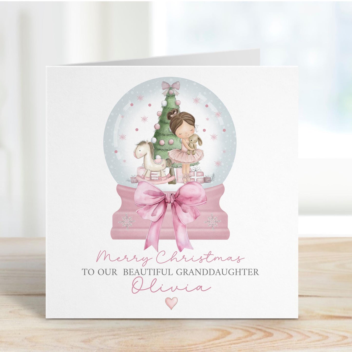 Square personalised Christmas card featuring a ballerina holding a teddy inside a pink snow globe with a Christmas tree, rocking horse, and presents. Text reads “Merry Christmas to our beautiful granddaughter/Daughter/Any relations&quot; Personalised with a name.