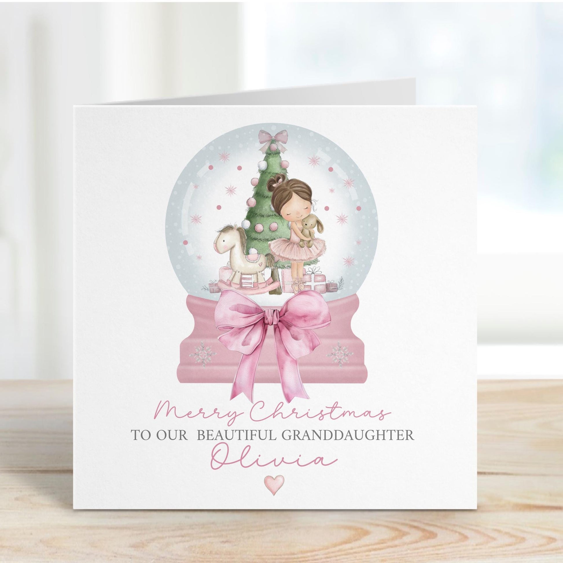 Square personalised Christmas card featuring a ballerina holding a teddy inside a pink snow globe with a Christmas tree, rocking horse, and presents. Text reads “Merry Christmas to our beautiful granddaughter/Daughter/Any relations&quot; Personalised with a name.