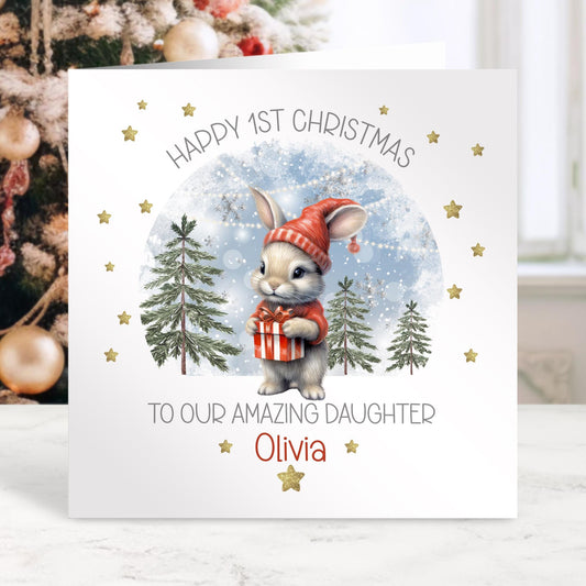 Cute first Christmas card featuring a cute bunny wearing a red hat & jumper holding a gift with a snowy background, surrounded by gold stars & the wording 'Happy 1st Christmas@ To our amazing daughter/Granddaughter/any relation, personalised with a name.
