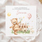 Square personalised second birthday greeting card featuring a cute illustrated teddy bear standing in a garden scene with flowers, a wooden fence, sunshine, gifts and a balloon showing the number two. The card includes personalised text for a child’s name and relation, with a soft pastel colour theme.